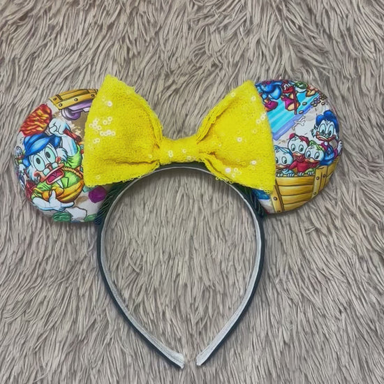 A headband featuring a colorful cartoon print of famous cartoon Duck characters, accented by a large yellow sequined bow in the center.