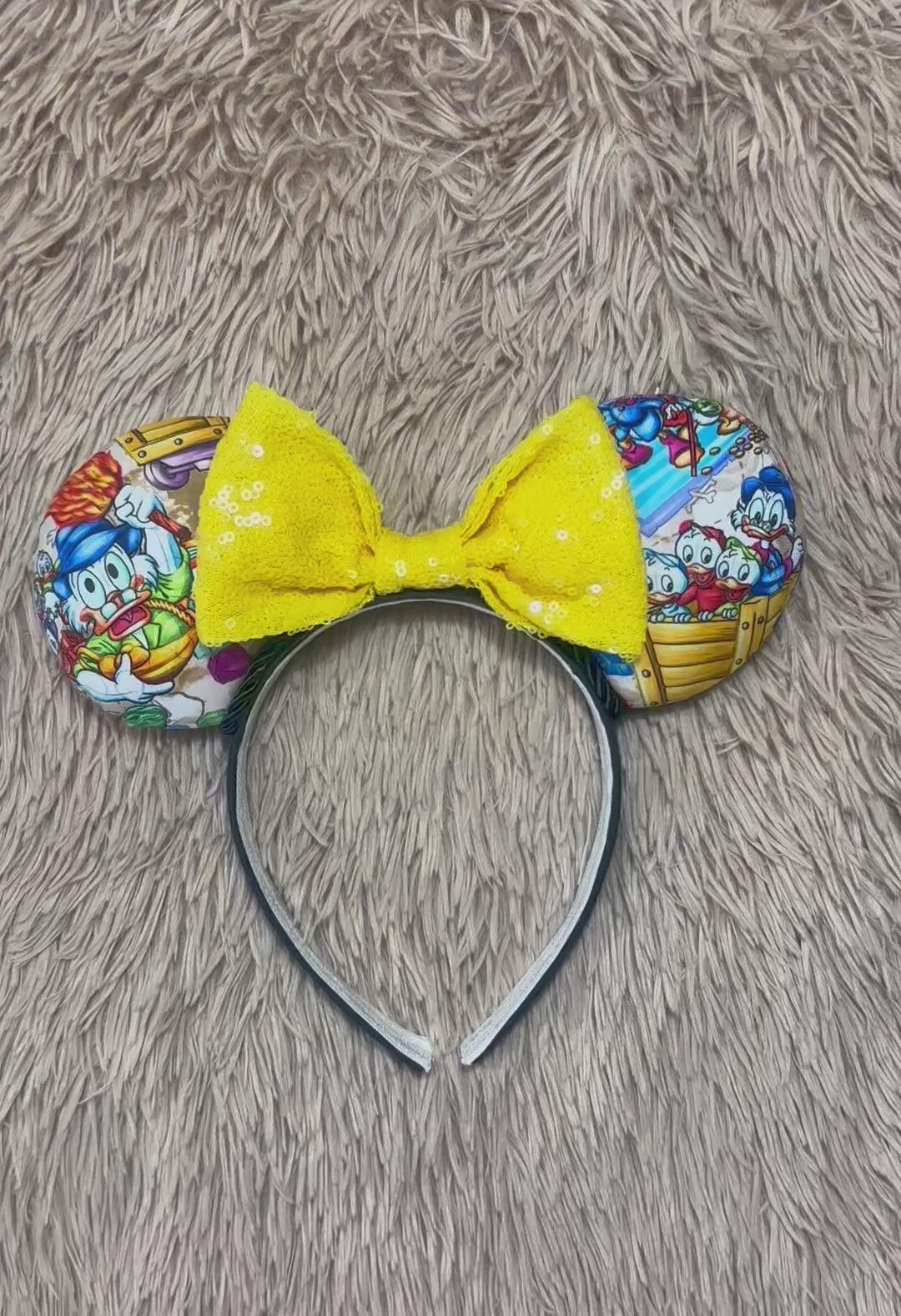 A headband featuring a colorful cartoon print of famous cartoon Duck characters, accented by a large yellow sequined bow in the center.
