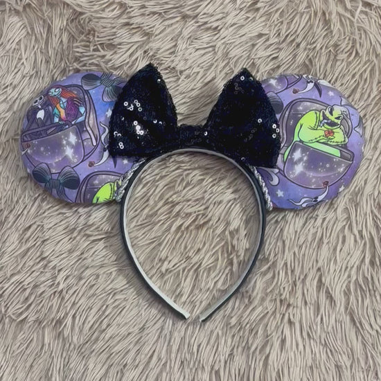 An ear headband accessory with a black sequined bow, featuring a purple HM and NBC themed print with a cartoon character in a buggy.