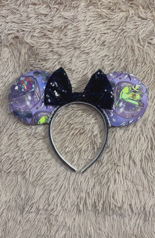 An ear headband accessory with a black sequined bow, featuring a purple HM and NBC themed print with a cartoon character in a buggy.