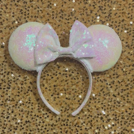 White Iridescent mouse ears with the fabric on both the front and back. The fabric is on the headband as well and is universal sized and fits most. The set has a matching iridescent sequin bow. Item is shown on a gold sequin background.