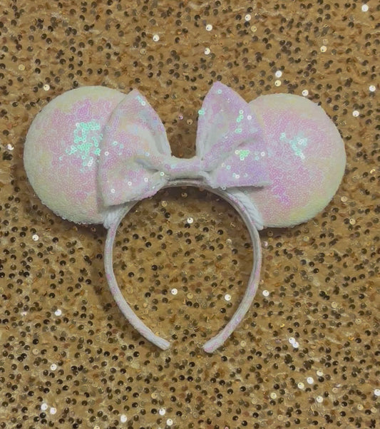 White Iridescent mouse ears with the fabric on both the front and back. The fabric is on the headband as well and is universal sized and fits most. The set has a matching iridescent sequin bow. Item is shown on a gold sequin background.
