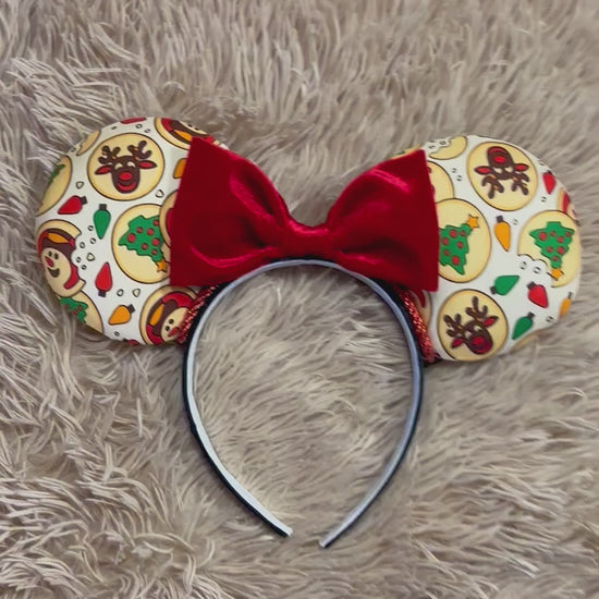 An ear headband featuring a festive Christmas print with red velvet bows, displayed on a beige faux fur background.