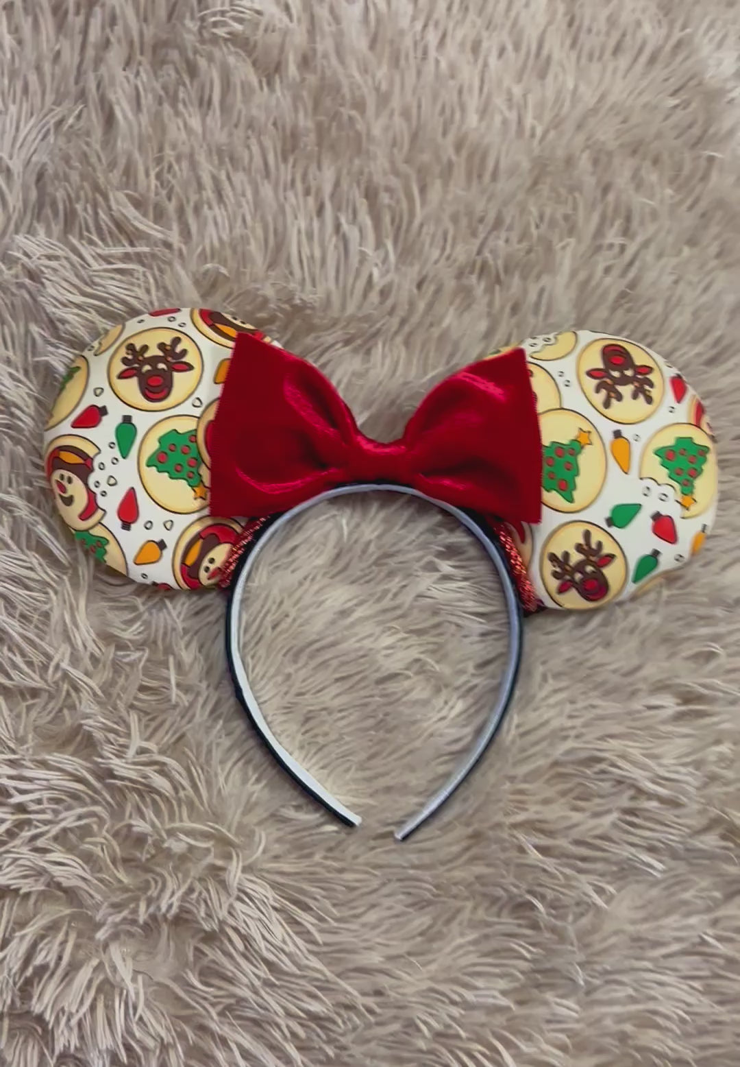 An ear headband featuring a festive Christmas print with red velvet bows, displayed on a beige faux fur background.