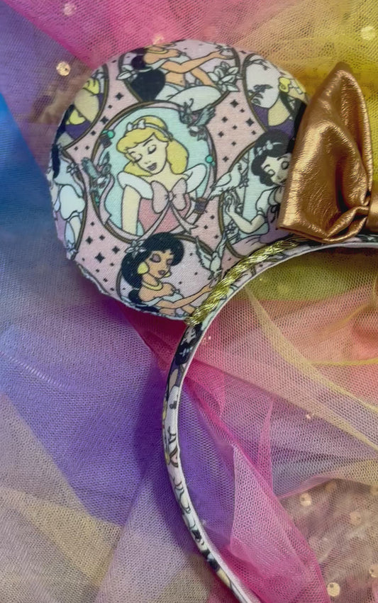 Headband with delicate blushing princesses on the ears and headband with a rose gold pleather bow in the middle. Shown on a rainbow background