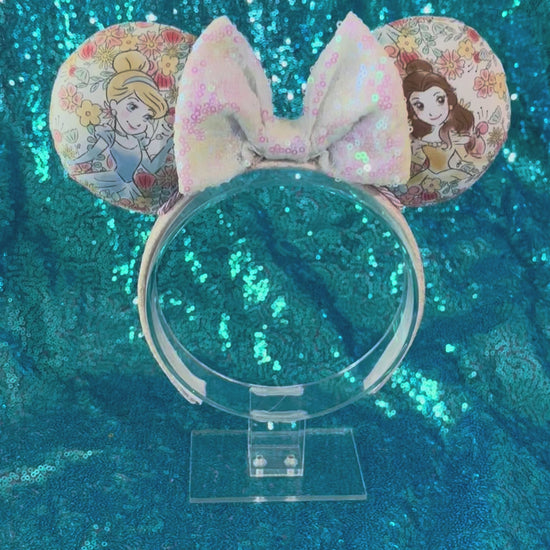 Handmade and ready to ship Pretty Princess Mouse ears with Cinderella and Belle shown on the fabric with a white iridescent bow. The headband has a white shimmery fabric and is a universal sized headband.