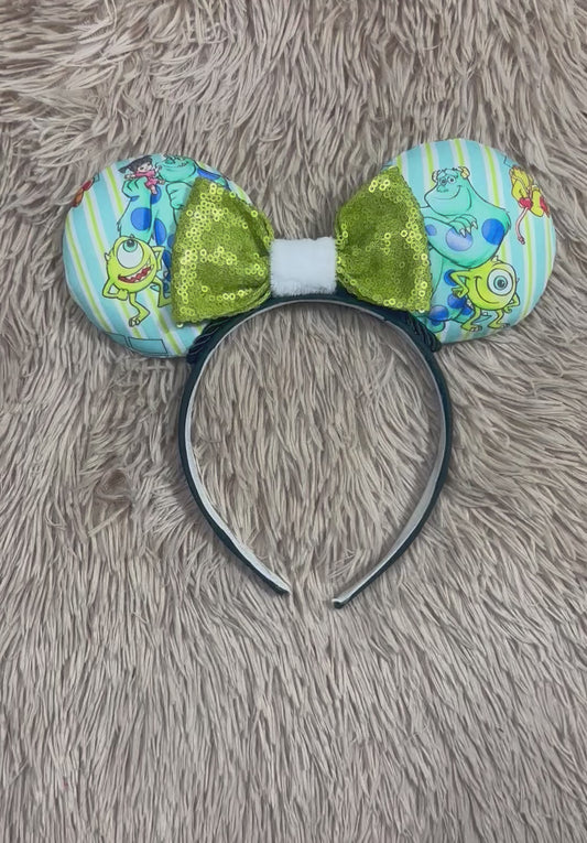 A Monster themed headband with two round ears featuring a light blue and yellow striped pattern with Mike and Sulley prints, accented by a large shiny olive green bow in the center and a white faux-fur trim.