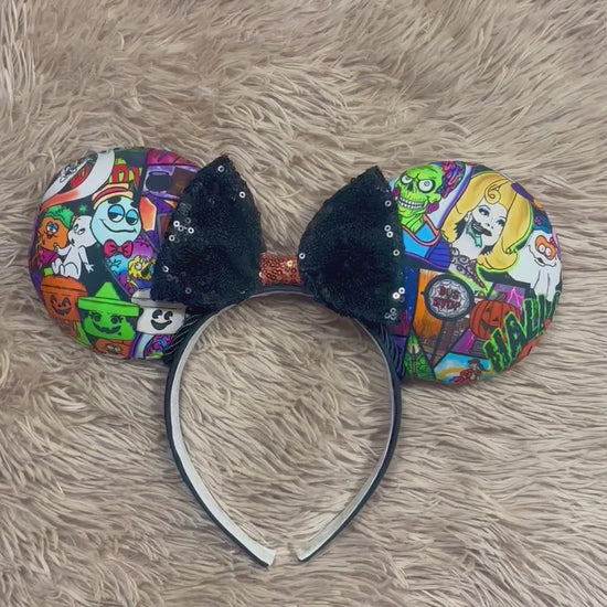 An ear headband accessory featuring a colorful Halloween-themed print on the outer ears and a black glitter bow in the center with black twisted trim along the inner ears.