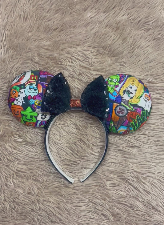 An ear headband accessory featuring a colorful Halloween-themed print on the outer ears and a black glitter bow in the center with black twisted trim along the inner ears.
