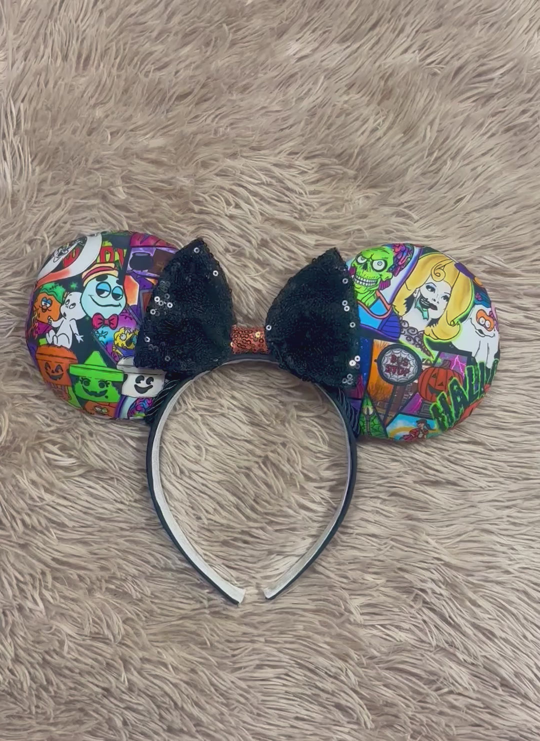 An ear headband accessory featuring a colorful Halloween-themed print on the outer ears and a black glitter bow in the center with black twisted trim along the inner ears.